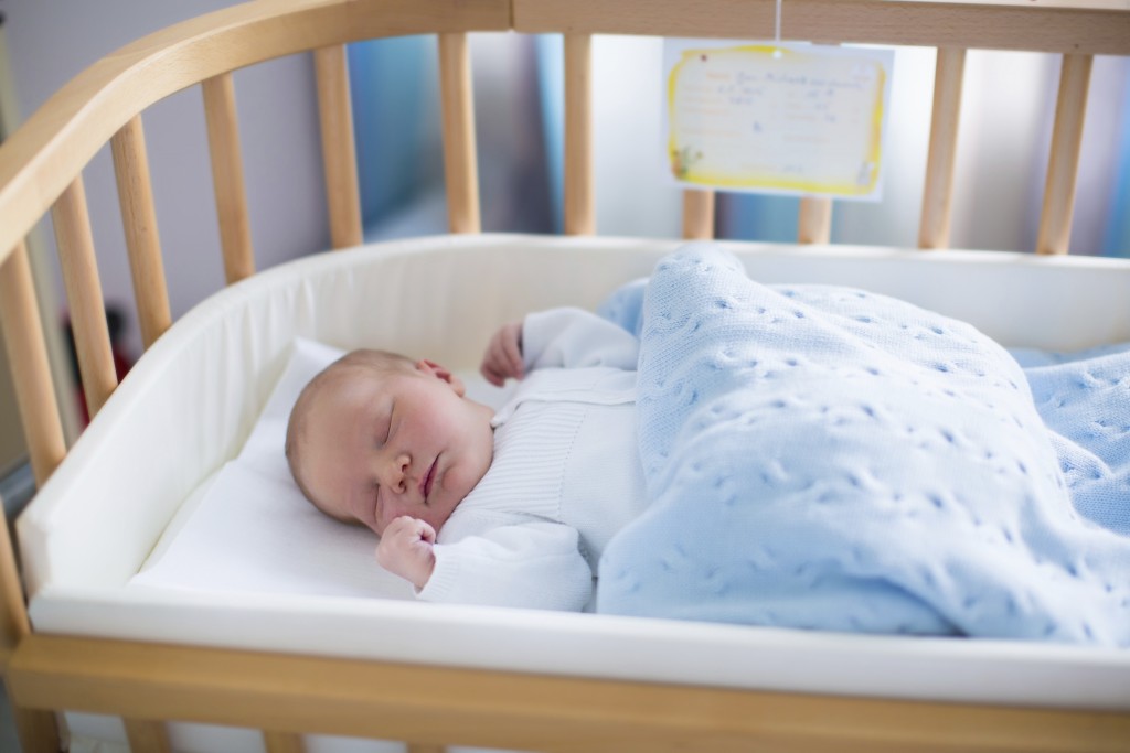 Newborn baby in hospital room. New born child in wooden co-sleeper crib. Infant sleeping in bedside bassinet. Safe co-sleeping in a bed side cot. Little boy taking a nap under knitted blanket.